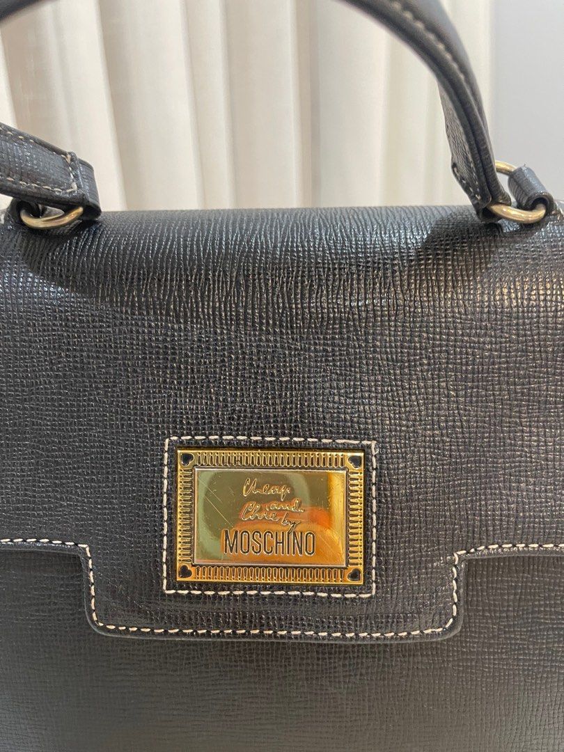 Vintage Moschino Black Handbag (Category: Luxury Bags & Wallets) located in Makati City. Submitted by triftedandbranded (Listing ID 1399383786, Image 2). Description: - Moschino black handbag - Top handle - Flap closure with gold-tone logo plaque - Interior lining with repeating Moschino logo pattern - White stitching details - Made of textured leather-like material.