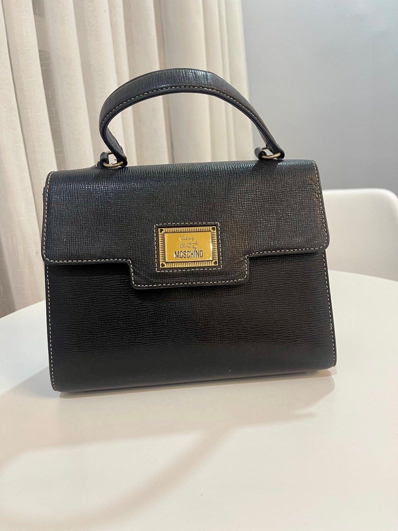 Vintage Moschino Black Handbag (Category: Luxury Bags & Wallets) located in Makati City. Submitted by triftedandbranded (Listing ID 1399383786, Image 1). Description: - Moschino black handbag - Top handle - Flap closure with gold-tone logo plaque - Interior lining with repeating Moschino logo pattern - White stitching details - Made of textured leather-like material.