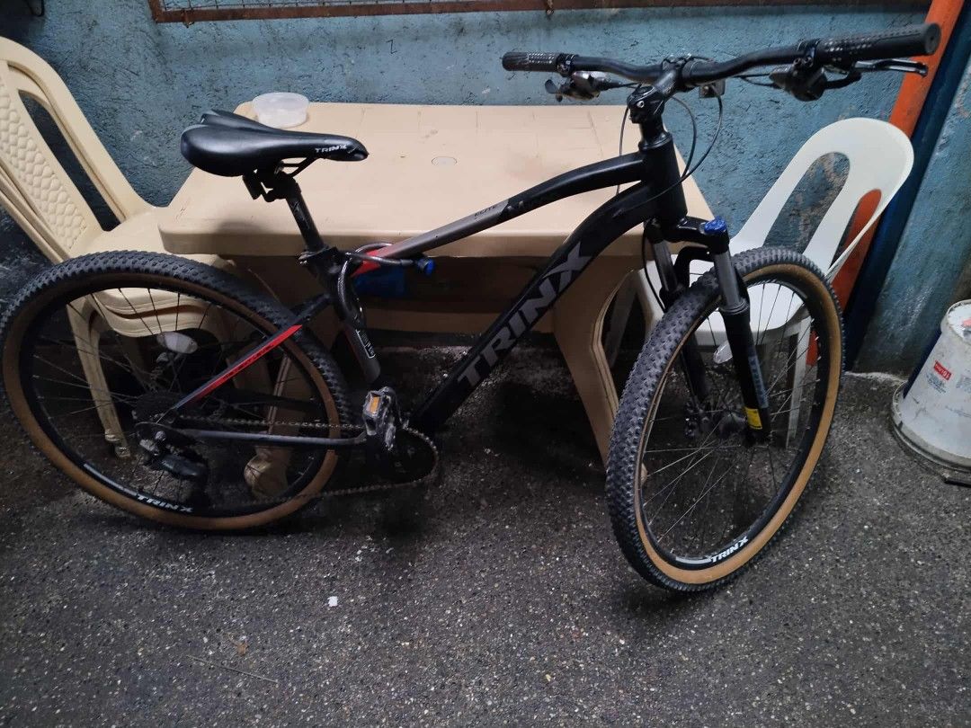 Trinx Mountain Bike (Category: Bicycles) located in Manila. Submitted by siredzelsa30772 (Listing ID 1400267505, Image 1). Description: Black Trinx mountain bike with tan tires. Features front suspension and disc brakes. The bike has a black frame with red accents. Includes a black seat and handlebars..