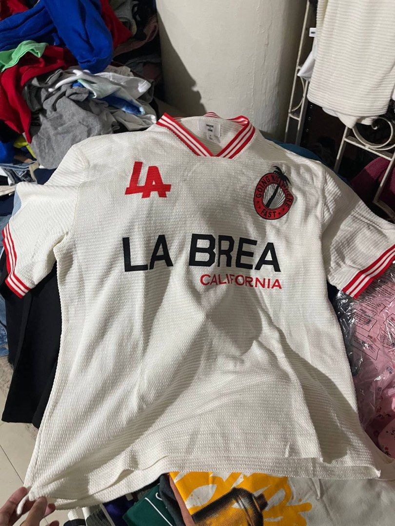 Mode of One La Brea Jersey (Category: Tshirts & Polo Shirts ) located in Obando. Submitted by Wintermelonzzzz (Listing ID 1401039071, Image 2). Description: Mode of One La Brea jersey. Features a white base with red accents on the collar and sleeves. The front displays "LA BREA CALIFORNIA" in bold black lettering, with "LA" on the upper left. A circular graphic with a palm tree and the words "GOING NOWHERE FAST" is on the right side. Made in Viet