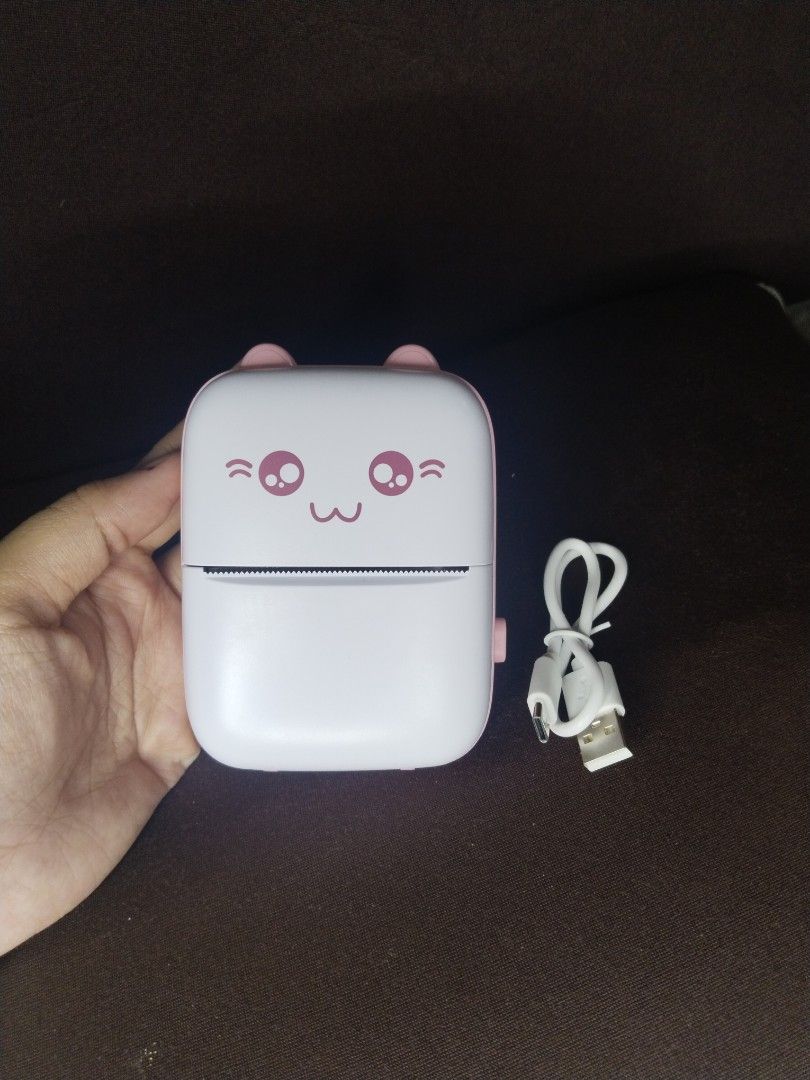 Portable Mini Printer - Cute Design (Category: Printers, Scanners & Copiers) located in San Luis. Submitted by thrift.with.shey (Listing ID 1401057669, Image 3). Description: Portable mini printer features a cute design. It comes with a charging cable and is packaged in its original box. The printer has a white and pink color scheme. The design includes a smiling face with cat ears..