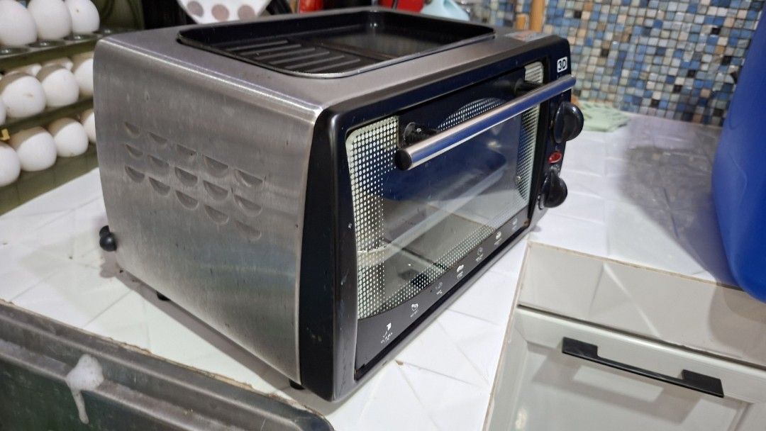 3D Toaster Oven with Grill (Category: Ovens & Toasters) located in Taguig. Submitted by oishibrownshishi (Listing ID 1401514713, Image 2). Description: 3D toaster oven features a grill on top and a glass door. It has two control knobs and a red button. The oven includes a baking tray and a wire rack. The exterior is stainless steel with black accents. It has a 3D logo on the front..