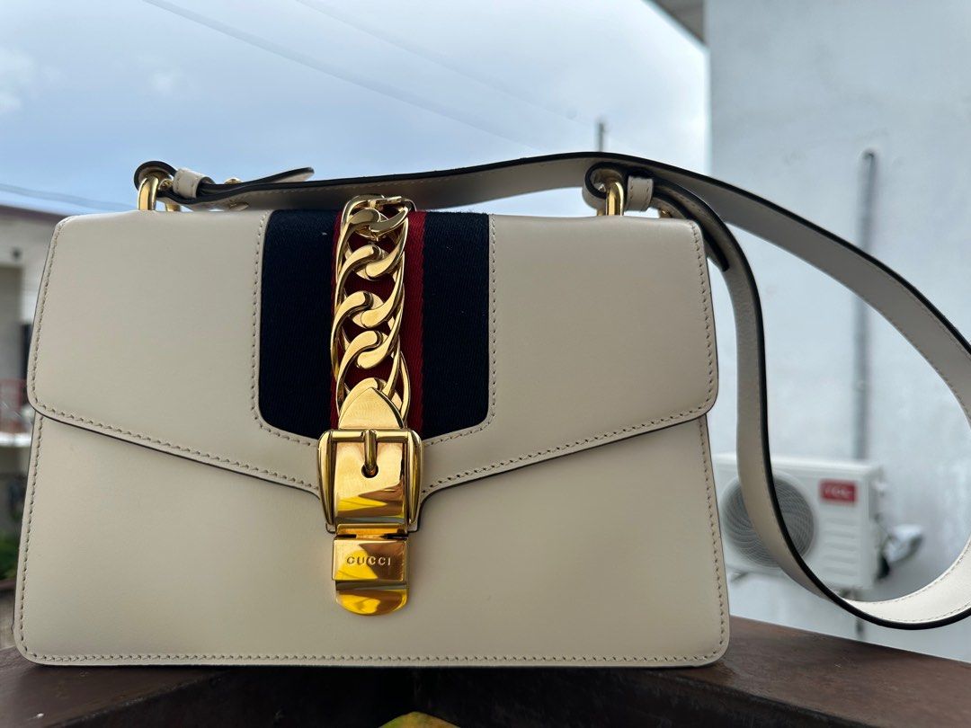 Gucci Sylvie Small Shoulder Bag (Authenticated by LegitGrails) (Category: Luxury Bags & Wallets) located in Manila. Submitted by kristineliban (Listing ID 1403645572, Image 2). Description: RFS: Need funds for home construction project With certificate of authenticity from LegitGrails Gucci Sylvie Small Shoulder Bag features a cream-colored leather exterior with a gold-tone chain and buckle closure. It has a signature red and navy stripe detail o