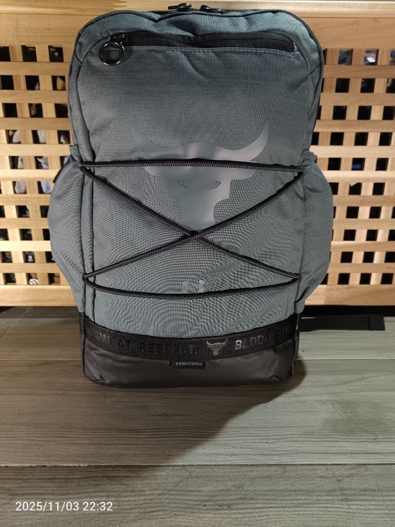 Under Armour Project Rock Brahma Backpack ( Gray ) (Category: Backpacks) located in Bacoor. Submitted by the.full.cart (Listing ID 1404723272, Image 1). Description: Under Armour Project Rock backpack features a gray color with the Project Rock logo. It has multiple compartments, including a front zippered pocket and side pockets. The backpack has adjustable straps and a top handle for easy carrying. The design includes black accents and the word