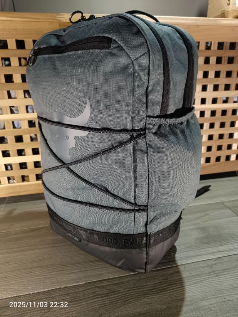 Under Armour Project Rock Brahma Backpack ( Gray ) (Category: Backpacks) located in Bacoor. Submitted by the.full.cart (Listing ID 1404723272, Image 2). Description: Under Armour Project Rock backpack features a gray color with the Project Rock logo. It has multiple compartments, including a front zippered pocket and side pockets. The backpack has adjustable straps and a top handle for easy carrying. The design includes black accents and the word