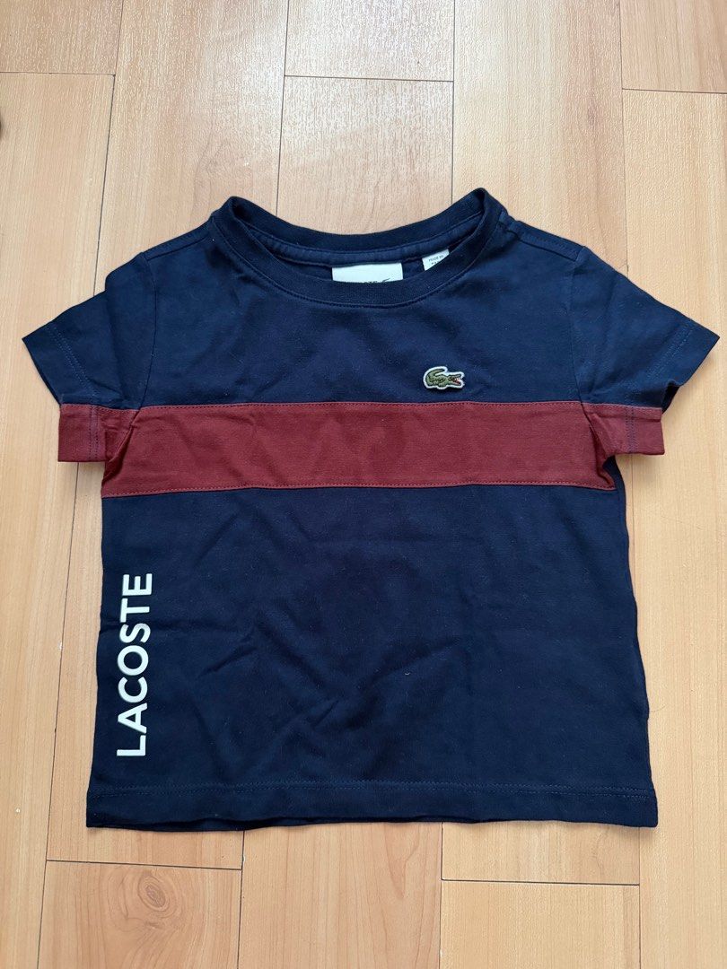 Lacoste Navy Blue T-shirt (Category: Babies & Kids Fashion) located in Manila. Submitted by ziaaa012 (Listing ID 1404964904, Image 1). Description: Lacoste T-shirt is navy blue with a burgundy stripe across the chest. It features the Lacoste logo on the chest and the brand name printed vertically on the side. The size is indicated as 2, equivalent to 92cm or 36 inches. Made in Peru. The fabric composition is 100% cotton..