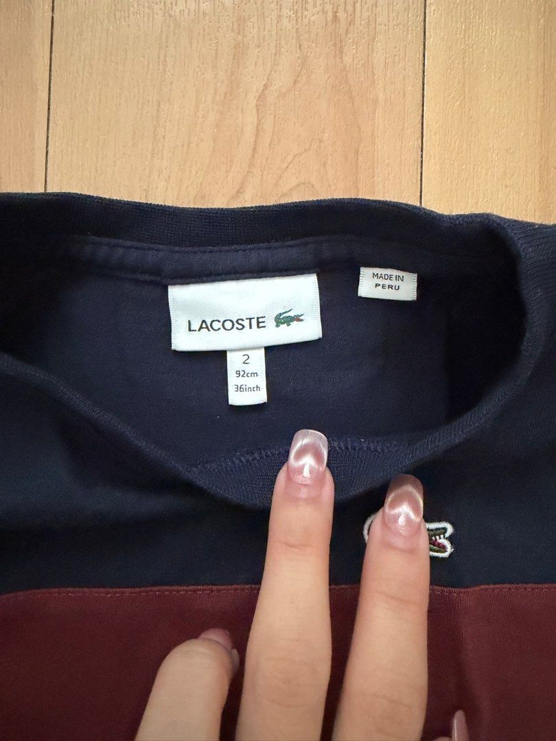 Lacoste Navy Blue T-shirt (Category: Babies & Kids Fashion) located in Manila. Submitted by ziaaa012 (Listing ID 1404964904, Image 2). Description: Lacoste T-shirt is navy blue with a burgundy stripe across the chest. It features the Lacoste logo on the chest and the brand name printed vertically on the side. The size is indicated as 2, equivalent to 92cm or 36 inches. Made in Peru. The fabric composition is 100% cotton..
