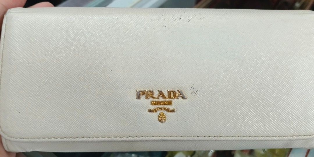 Prada Long Wallet (Category: Luxury Bags & Wallets) located in Legazpi City. Submitted by georgepineda791 (Listing ID 1401717114, Image 1). Description: Preloved Prada Long Wallet in light cream color. Features two-button lock with Prada engraving, multiple card slots, coin slot, etiketa, and code inside. Flaws: rubs on edges and body, fading hardware, tiny cracks on edges, yellowish glue stain inside pocket, and dirt/stain on the side part (poss
