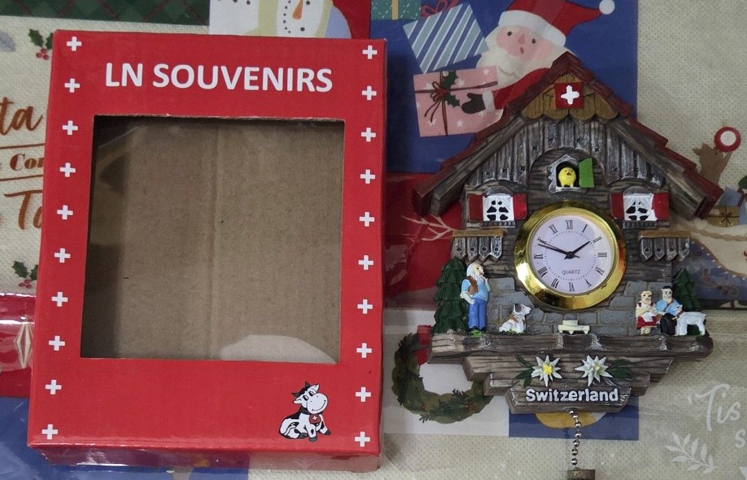 LN Souvenirs Switzerland Cuckoo Clock Magnet (Category: Clocks) located in Manila. Submitted by sterling_grace (Listing ID 1406049259, Image 1). Description: Charming cuckoo clock is from LN Souvenirs and features a Swiss design. It has a wooden house shape with a clock face and decorative figurines. The clock is in its original box, which is red with white crosses and a cow illustration. The box is labeled "LN Souvenirs", bought in Switzerland..