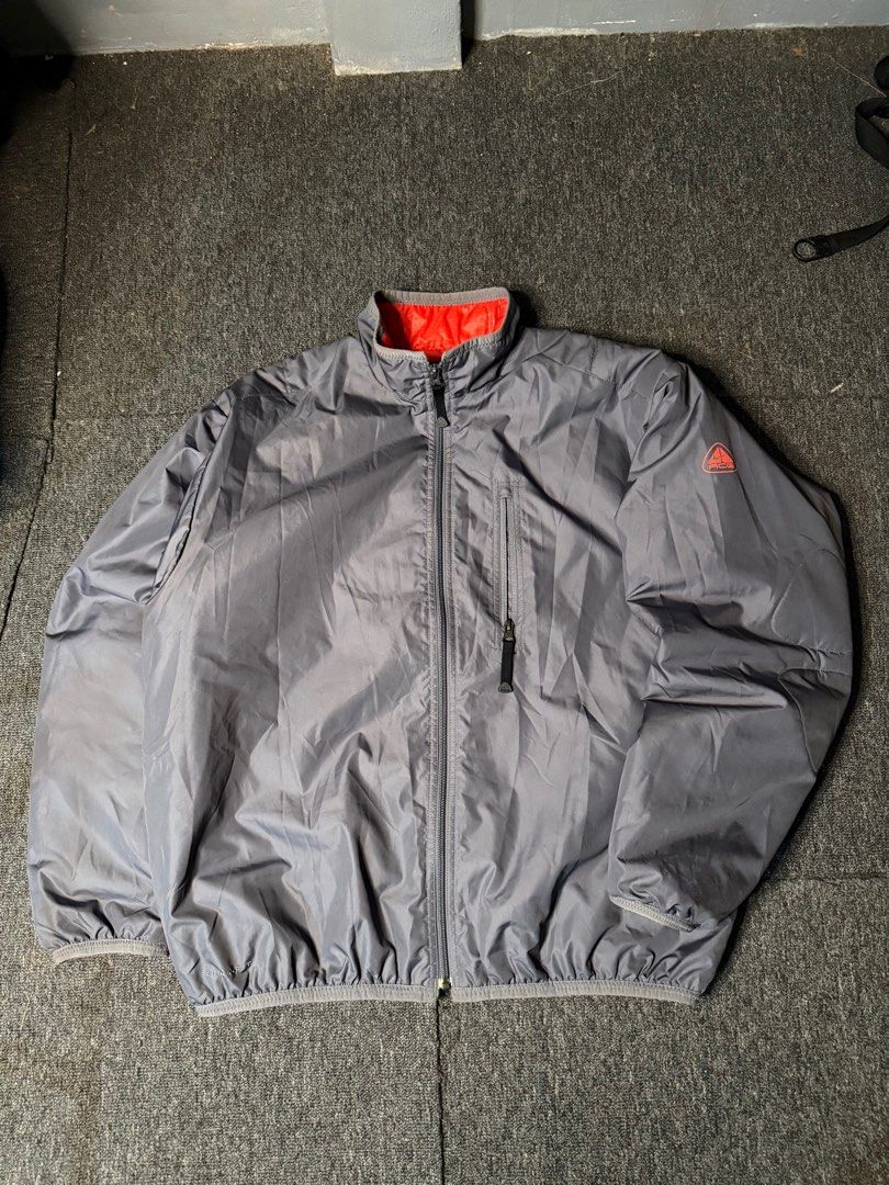 Nike ACG Puffer Windbreaker (Category: Coats, Jackets and Outerwear) located in San Marcelino. Submitted by SoloMain (Listing ID 1406055118, Image 1). Description: Nike ACG puffer windbreaker features a gray exterior with a red interior. It has a zip closure, a pocket on the chest, and the ACG logo on the sleeve. The jacket is labeled as Clima-Fit. The width is 25.5 inches and the length is 27.5 inches..