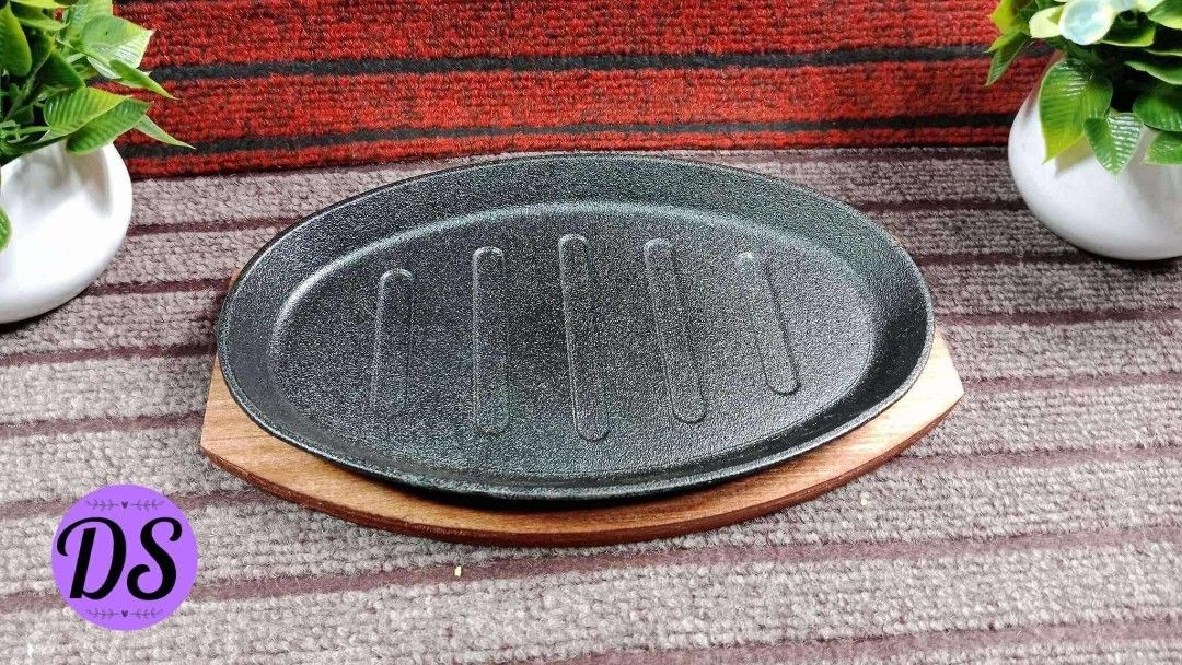 Oval Sizzling Plate with Wooden Base (Category: Cookware & Accessories) located in Mandaluyong City. Submitted by shopmla (Listing ID 1402191951, Image 3). Description: Oval sizzling plate with a wooden base. The plate has a black, textured surface with grooves. The wooden base is reddish-brown and shaped to fit the plate. Perfect for serving hot dishes. Great for restaurants or home use..