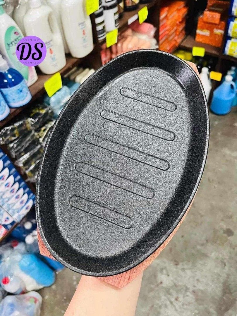 Oval Sizzling Plate with Wooden Base (Category: Cookware & Accessories) located in Mandaluyong City. Submitted by shopmla (Listing ID 1402191951, Image 2). Description: Oval sizzling plate with a wooden base. The plate has a black, textured surface with grooves. The wooden base is reddish-brown and shaped to fit the plate. Perfect for serving hot dishes. Great for restaurants or home use..