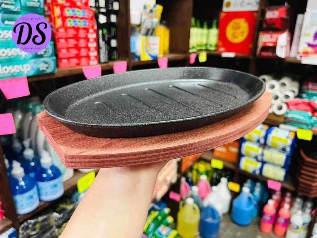 Oval Sizzling Plate with Wooden Base (Category: Cookware & Accessories) located in Mandaluyong City. Submitted by shopmla (Listing ID 1402191951, Image 1). Description: Oval sizzling plate with a wooden base. The plate has a black, textured surface with grooves. The wooden base is reddish-brown and shaped to fit the plate. Perfect for serving hot dishes. Great for restaurants or home use..