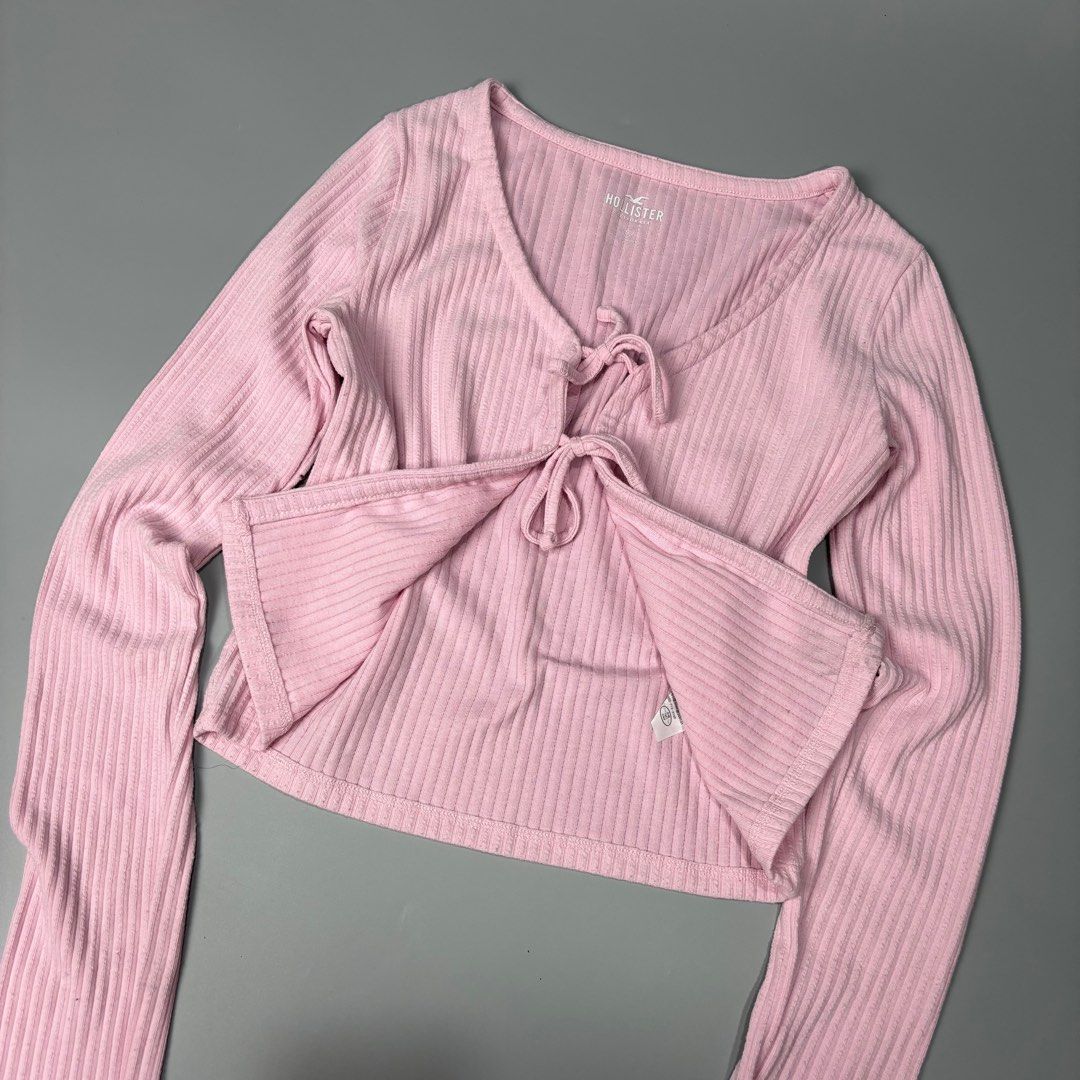 Hollister Pink Ribbed Tie-Front Top (Category: Sleeveless) located in Angeles City. Submitted by tey68 (Listing ID 1402802822, Image 2). Description: Hollister top features a soft pink color and a ribbed texture. It has long sleeves and a tie-front closure. The top is a cropped style. The Hollister logo is visible on the front. The size tag is also visible in the images..