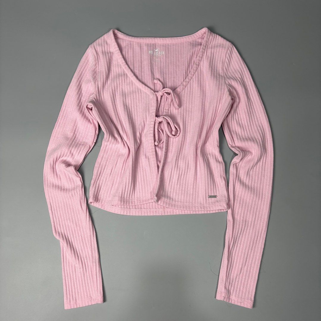 Hollister Pink Ribbed Tie-Front Top (Category: Sleeveless) located in Angeles City. Submitted by tey68 (Listing ID 1402802822, Image 1). Description: Hollister top features a soft pink color and a ribbed texture. It has long sleeves and a tie-front closure. The top is a cropped style. The Hollister logo is visible on the front. The size tag is also visible in the images..
