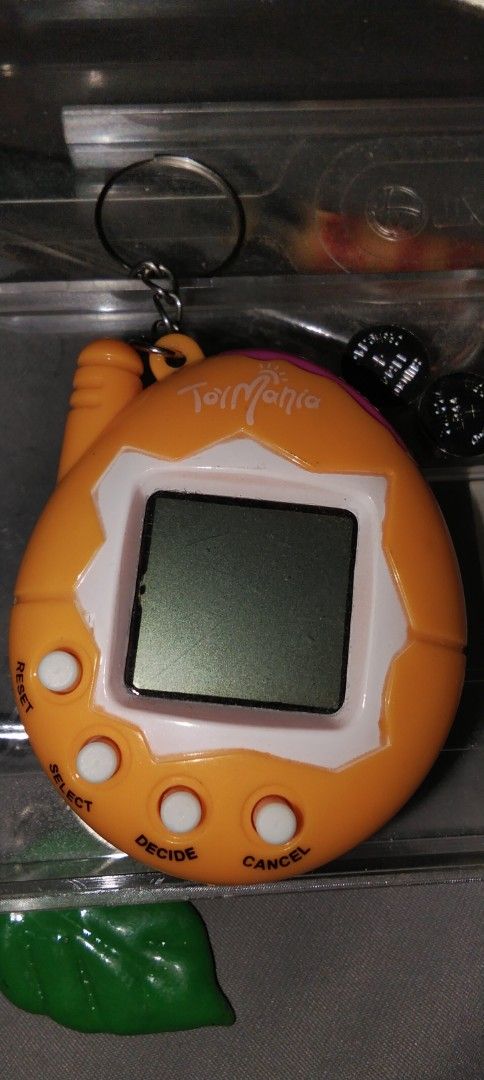 Toy Mania Virtual Pet (Category: Interactive Toys) located in Santa Rosa. Submitted by animemerchcollection (Listing ID 1409084291, Image 2). Description: - Tamagotchi Mania virtual pet - Orange and white design - Small screen and buttons - Pixelated character display - Working condition - Includes keychain attachment - Battery compartment at the back.