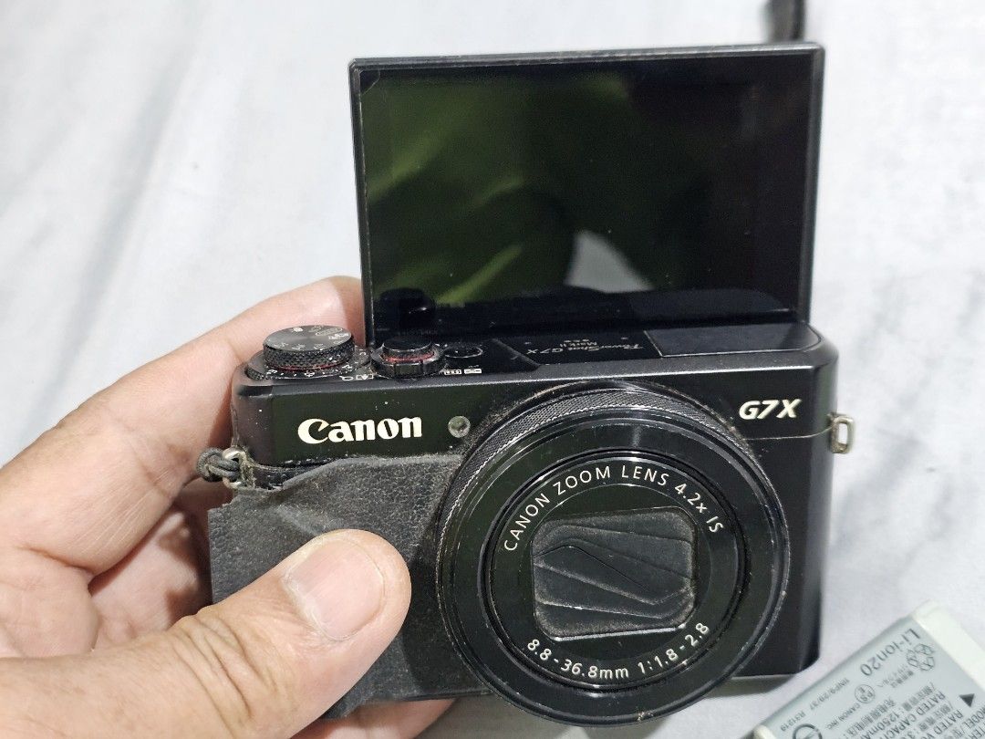 Canon PowerShot G7 X Mark II Digital Camera (Category: Cameras) located in Tagbilaran City. Submitted by lugarpogi (Listing ID 1409589463, Image 1). Description: Canon PowerShot G7 X Mark II digital camera. Includes the camera, three batteries, and a battery charger. Features a flip-up screen and a zoom lens. The camera has a strap attached. The screen displays various settings and captured images. The camera is black..