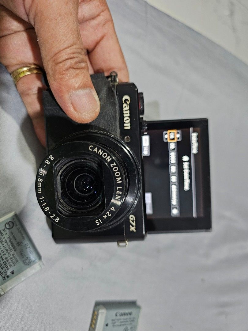 Canon PowerShot G7 X Mark II Digital Camera (Category: Cameras) located in Tagbilaran City. Submitted by lugarpogi (Listing ID 1409589463, Image 3). Description: Canon PowerShot G7 X Mark II digital camera. Includes the camera, three batteries, and a battery charger. Features a flip-up screen and a zoom lens. The camera has a strap attached. The screen displays various settings and captured images. The camera is black..