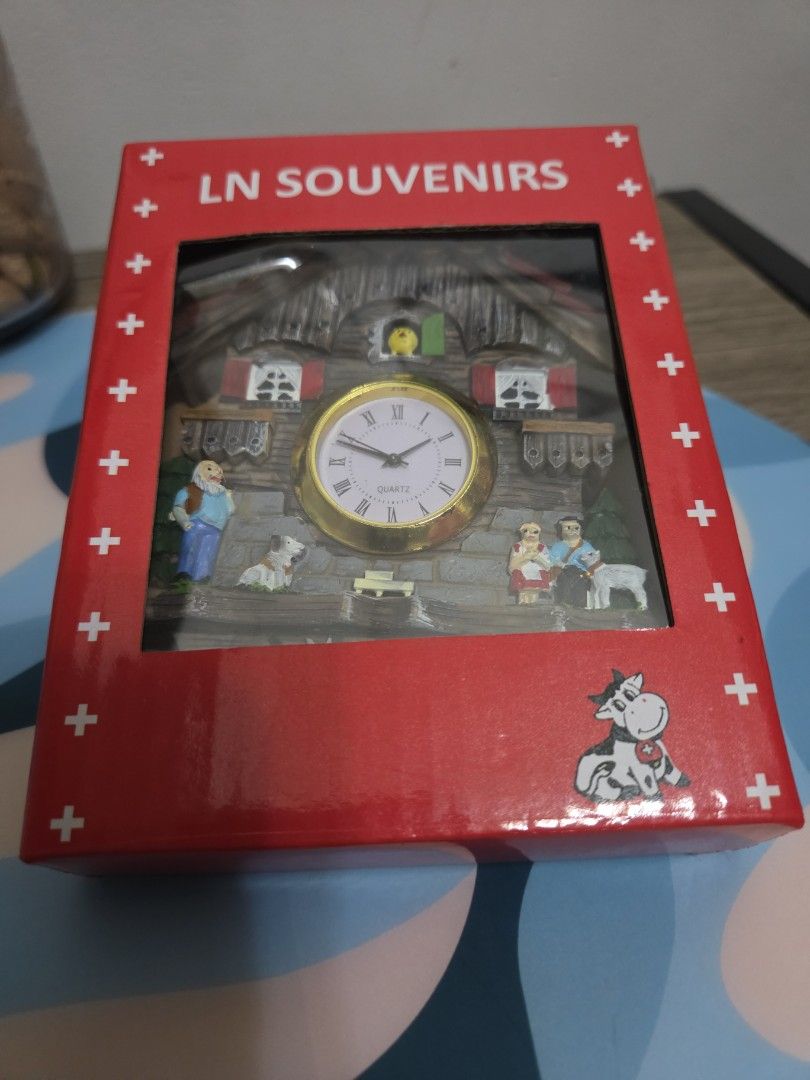 LN Souvenirs Switzerland Cuckoo Clock Magnet (Category: Clocks) located in Manila. Submitted by sterling_grace (Listing ID 1406049259, Image 2). Description: Charming cuckoo clock is from LN Souvenirs and features a Swiss design. It has a wooden house shape with a clock face and decorative figurines. The clock is in its original box, which is red with white crosses and a cow illustration. The box is labeled "LN Souvenirs", bought in Switzerland..
