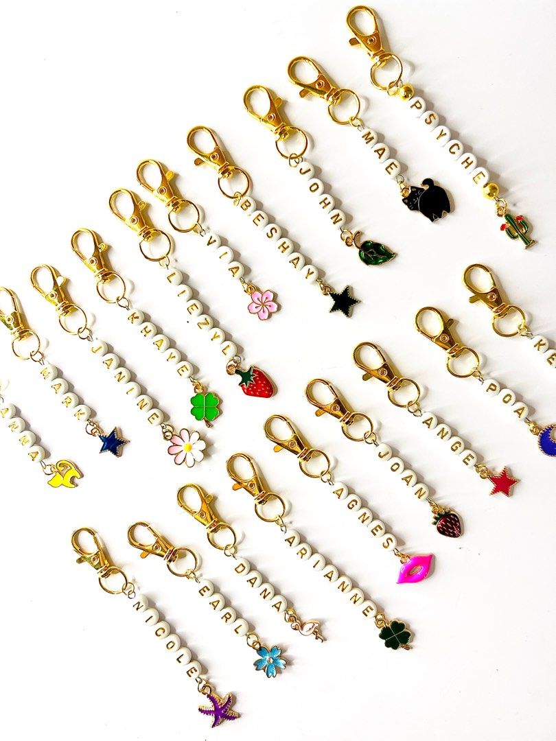 Personalized Name Keychains with Charms (Category: Charms) located in Bacoor. Submitted by MAB_Store (Listing ID 1407561391, Image 1). Description: Customizable keychains featuring names spelled out with letter beads. Each keychain includes a gold-colored clasp and a charm. Names visible include: Psyche, Mae, John, Beshay, Via, Agnes, Arianne, Dana, Mark, Earl, Anna, and Nicole. Charms include stars, flowers, a strawberry, a four-leaf clover, a l
