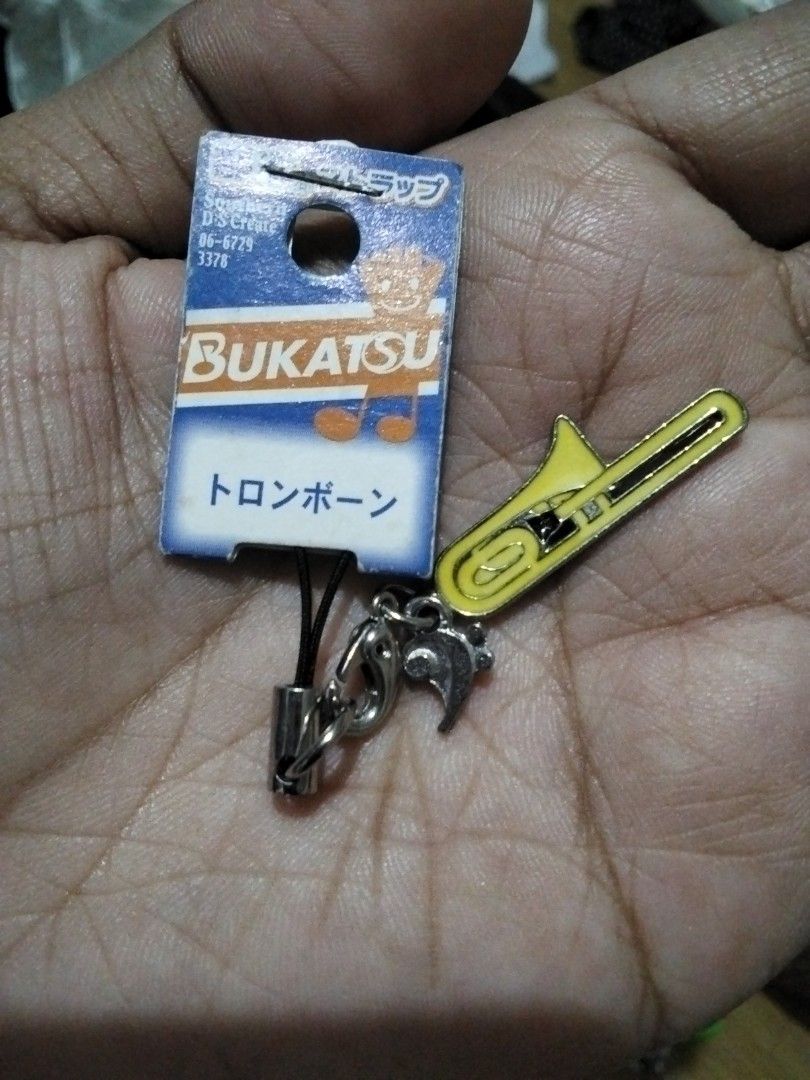 Bukatso Trombone Charm (Category: Charms) located in Santa Rosa. Submitted by animemerchcollection (Listing ID 1410822855, Image 1). Description: - Bukatso trombone charm - Yellow trombone design - Silver-colored chain - Black strap - Includes original packaging - Packaging features Japanese text and a bear illustration - Perfect for music enthusiasts - Unique and cute accessory.