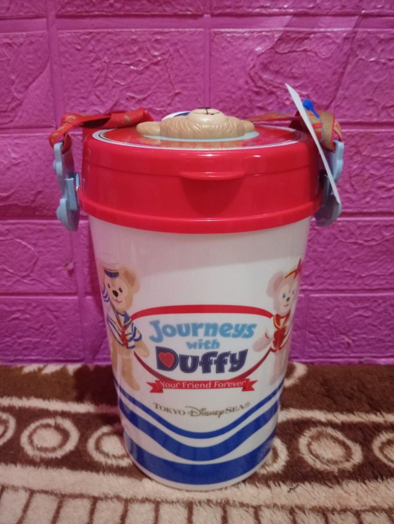 Duffy Journeys pop corn bucket (Category: Water Bottles & Tumblers) located in Bulacan. Submitted by renasophiatoystore2020 (Listing ID 1412215381, Image 1). Description: Tumbler features Duffy from Tokyo DisneySea's "Journeys with Duffy" collection. It has a red lid with a Duffy design and a matching strap. The tumbler is white with blue accents and Duffy graphics. It includes a tag with a price of ¥110. The tumbler is designed to keep your drin