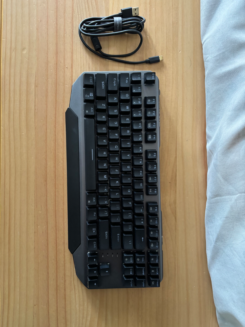 Machenike Mechanical Keyboard with Blue Backlight (Category: Computer Keyboard) located in Caloocan City. Submitted by bloomstoday (Listing ID 1408537010, Image 3). Description: Machenike - Mechanical keyboard - Blue backlight - Standard layout with function keys and number pad - Black keys with white lettering - Includes USB cable - Unique angled design - Shown on a wooden surface.