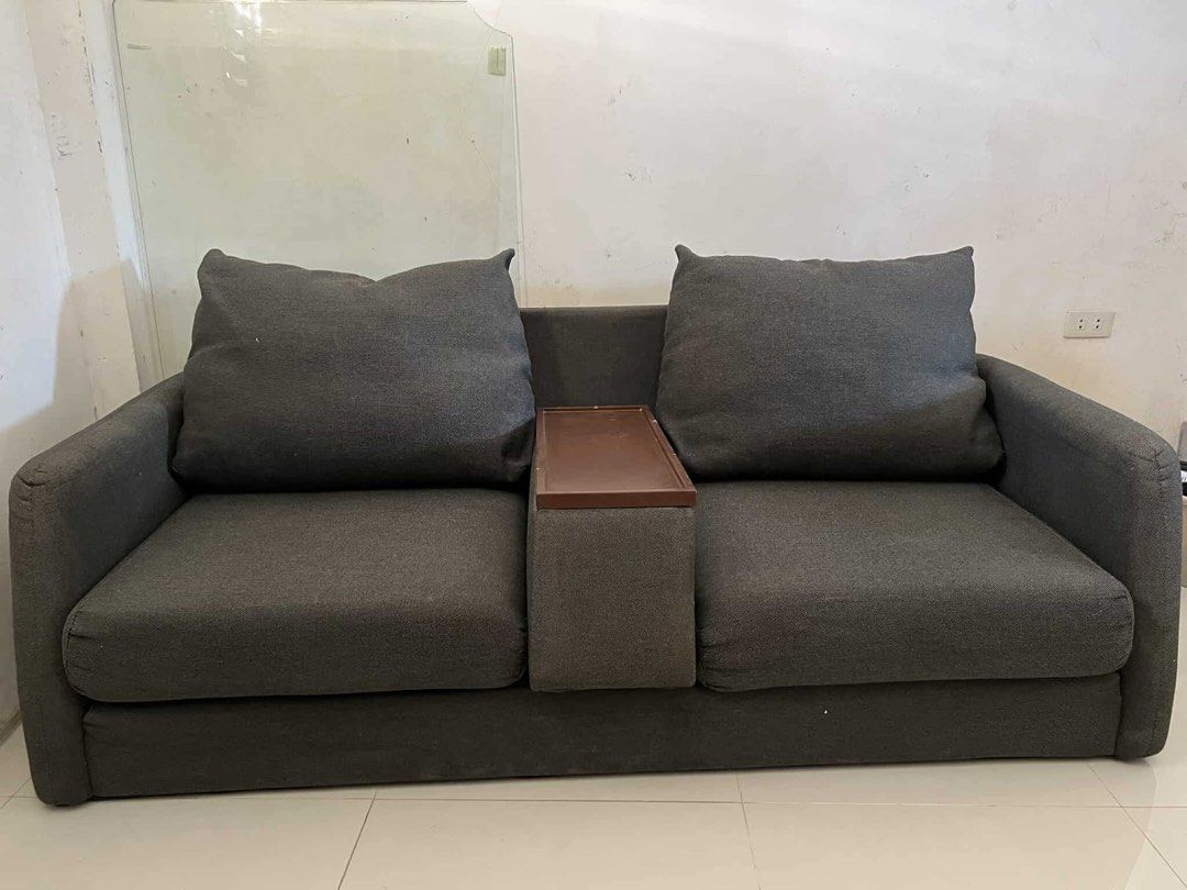 Sofa Japan Surplus (Category: Sofas) located in Makati City. Submitted by thiftdirection (Listing ID 1367388452, Image 1). Description: A compact, modern two-seater sofa with dark gray fabric upholstery. The design is minimalist, featuring straight arms and large, plush backrest cushions. A unique detail is the built-in tray or console between the two seats, likely for placing drinks or small items, enhancing its functionality for casual seating 