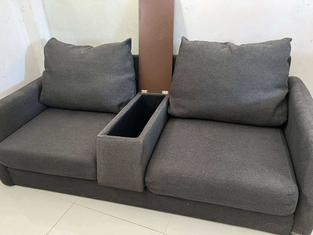 Sofa Japan Surplus (Category: Sofas) located in Makati City. Submitted by thiftdirection (Listing ID 1367388452, Image 3). Description: A compact, modern two-seater sofa with dark gray fabric upholstery. The design is minimalist, featuring straight arms and large, plush backrest cushions. A unique detail is the built-in tray or console between the two seats, likely for placing drinks or small items, enhancing its functionality for casual seating 