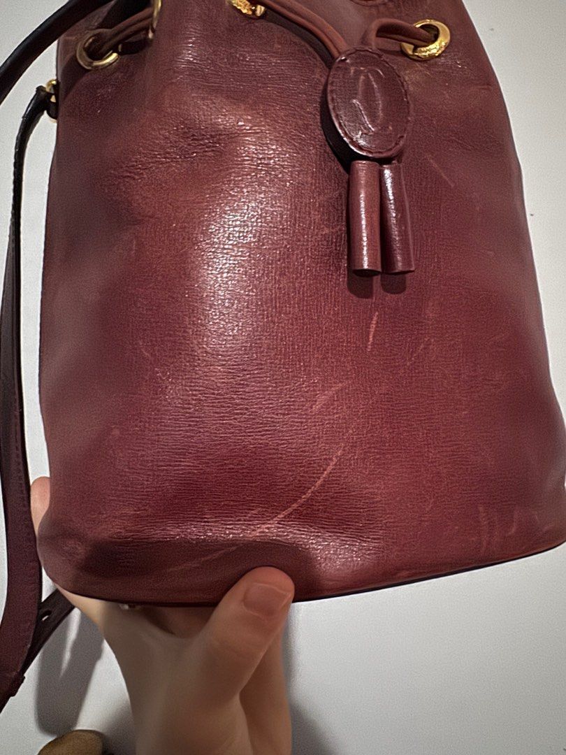 Cartier Bucket Bag - Burgundy (Category: Bags & Wallets) located in Singapore. Submitted by aroundtheworld19 (Listing ID 1378786514, Image 3). Description: Authentic Cartier bucket bag in burgundy leather. Features a drawstring closure, gold-tone hardware, and an adjustable shoulder strap. Interior includes a small zip pocket. Shows signs of wear, including scratches on the exterior. A classic and stylish piece. Note condition on one side of the 
