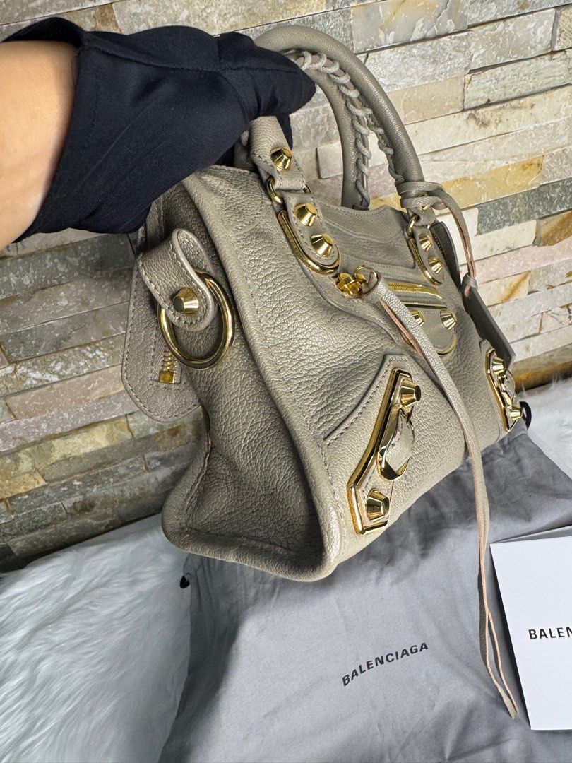 Balenciaga Mini City (Category: Luxury Bags & Wallets) located in Taguig. Submitted by angelbellaclaire (Listing ID 1386466508, Image 2). Description: Balenciaga City Bag – Mini – Taupe with Gold Hardware • Material: Premium grained lambskin leather • Color: Taupe (neutral gray-brown tone) • Hardware: Shiny gold-tone studs and zipper pulls • Features: Signature front zipper pocket, detachable leather-framed mirror, top zip closure, dual rolled le