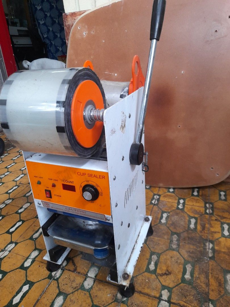 Cup Sealer Machine (Category: Other Kitchen Appliances) located in Valenzuela. Submitted by newbear.425b6e (Listing ID 1394877344, Image 2). Description: - Cup sealer machine - Orange control panel with count display - Temperature control - Power switch - Includes handle for operation - Comes with a roll of sealing film - Designed for efficient cup sealing.