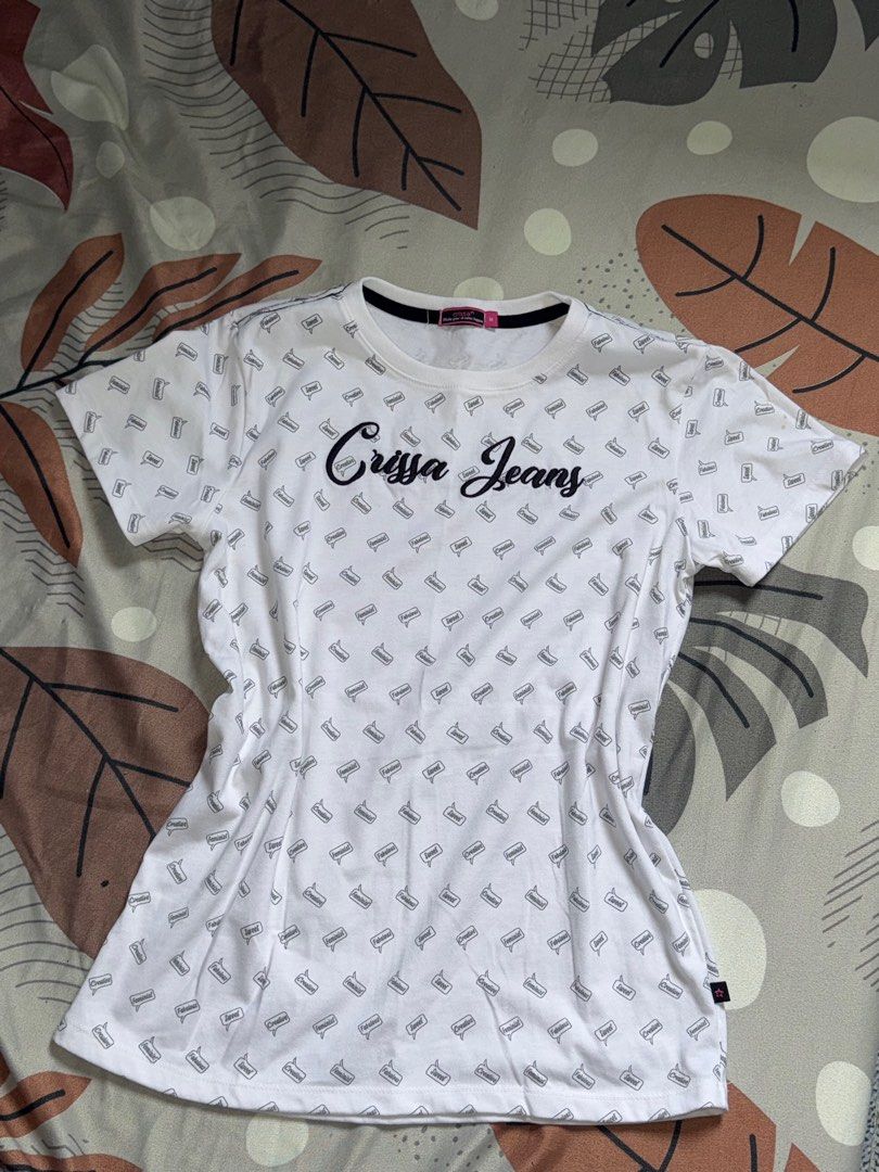 Crissa Jeans White T-shirt (Category: Shirts) located in Mandaluyong City. Submitted by quickriver.c73050 (Listing ID 1395015544, Image 1). Description: White Crissa Jeans t-shirt with a crew neck. Features a repeating pattern of small images and the brand name embroidered on the front. The tag indicates a size M. The shirt has short sleeves and a standard fit. The shirt is white with black accents..