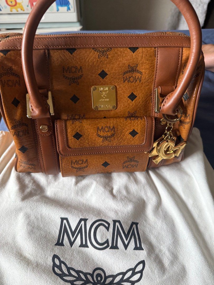 Authentic MCM Boston Bag with Charm (Category: Luxury Bags & Wallets) located in Manila. Submitted by grandeagle-f103ae (Listing ID 1414690152, Image 1). Description: - Authentic MCM Boston bag - Iconic MCM logo pattern (cognac) - Gold-tone hardware - Detachable charm - Top handle and detachable shoulder strap - Fabric lining with small pocket - Includes dust bag - Made in Korea -with free generic strap.