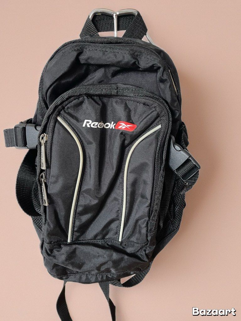 REEBOK BACKPACK (Category: Backpacks) located in San Fernando. Submitted by japansurpluselyu (Listing ID 1417021389, Image 1). Description: Dimes: 11x14 black Reebok mini backpack with a front zippered pocket featuring white trim and the “Reebok” logo in white letters alongside a red triangular emblem with a white Reebok vector. has adjustable shoulder straps, a top handle with a metal D-ring, and additional side pockets with plastic buckles..