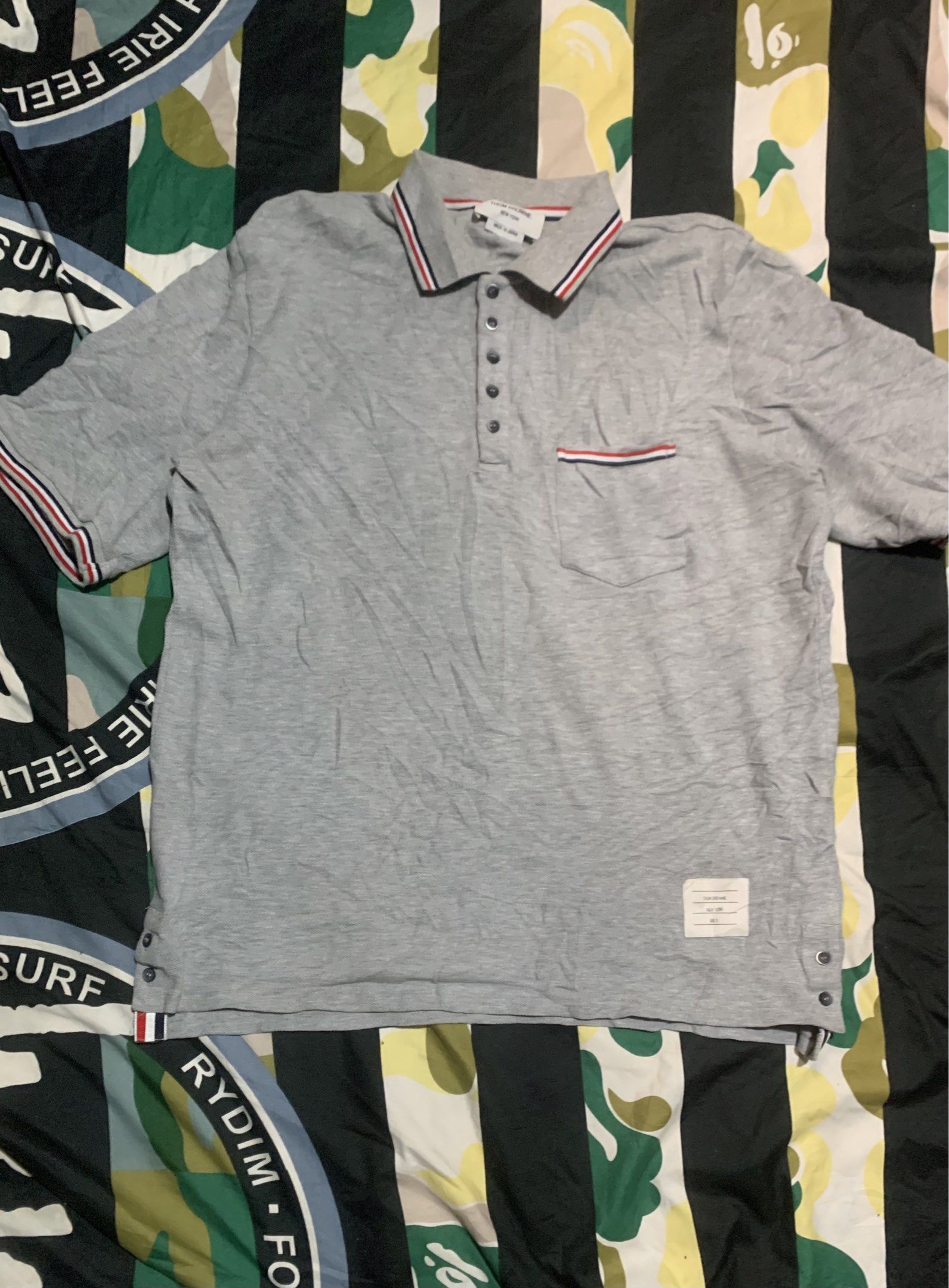 Thom Browne Grey Polo Shirt Size 3 (Category: Luxury Apparel) located in Cagayan de Oro. Submitted by agboysalis60090 (Listing ID 1417243075, Image 1). Description: Authentic Thom Browne polo shirt in grey. Features the signature red, white, and blue trim on the collar, pocket, and sleeves. Made in Japan. Size 3. Includes a chest pocket and button-up placket. The shirt has a classic polo design with short sleeves..