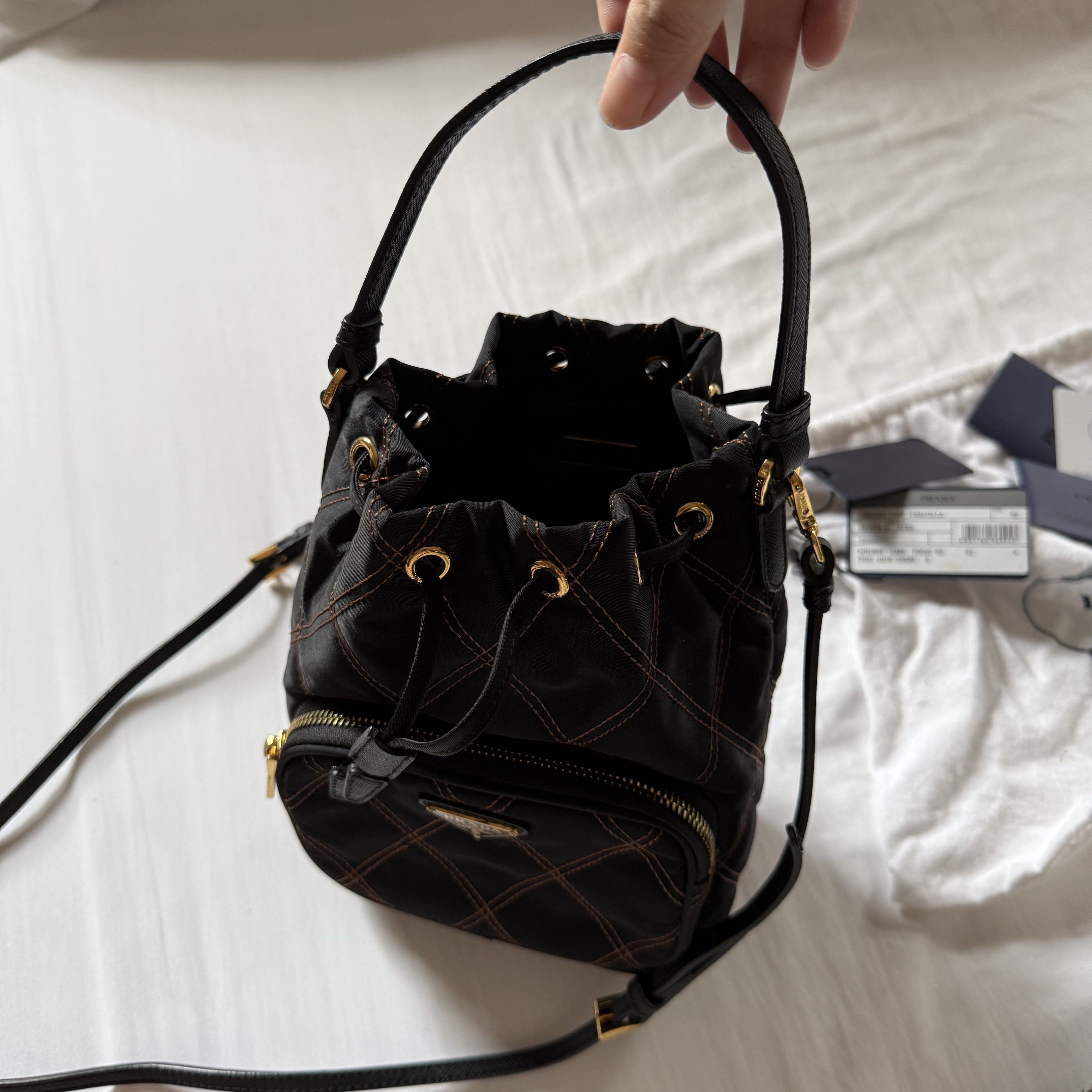 Prada Quilted Nylon Bucket Bag (Category: Luxury Bags & Wallets) located in Quezon City. Submitted by aligatsss (Listing ID 1417615498, Image 3). Description: - NGA Prada bucket bag - Black nylon with gold-tone hardware - Drawstring closure - Front pocket with Prada logo - Detachable shoulder strap - Includes authenticity cards and dust bag - Interior features Prada Milano logo - Quilted design with gold stitching.
