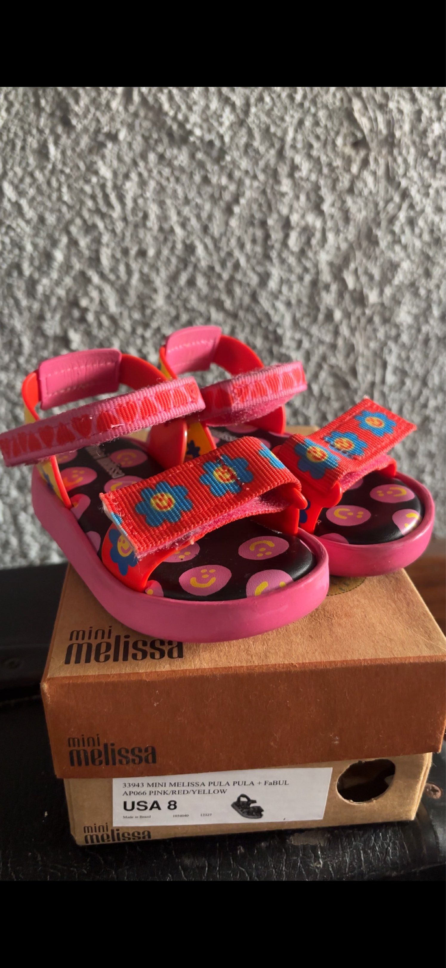 Mini Melissa Pula Pula Sandals 8C (Category: Babies & Kids Fashion) located in San Jose. Submitted by arakyer (Listing ID 1417822605, Image 1). Description: - Mini Melissa Pula Pula sandals - Available in pink, red, and yellow - Features a floral design on the straps - Smiley face pattern on the footbed - Includes original box - Size: USA 8.