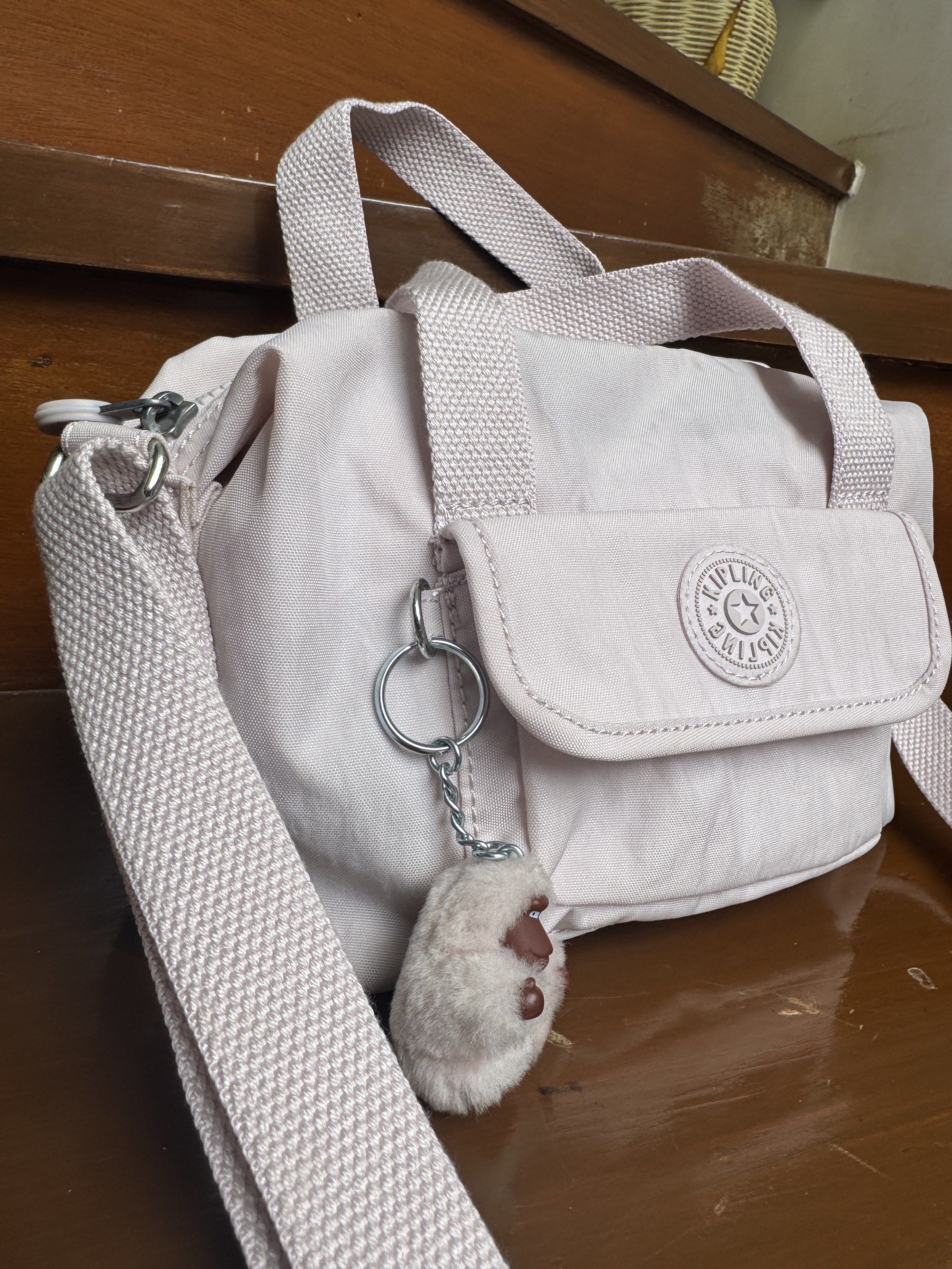 Kipling Brynn Serene Lavender Crossbody Bag (Category: Cross-body Bags) located in Paranaque City. Submitted by kenoh07 (Listing ID 1418185205, Image 2). Description: Kipling Brynn Serene Lavender crossbody bag. Features a light purple color with a detachable strap and top handles. Includes a small front pocket and a Kipling monkey keychain. The interior has a white lining with a small pocket. The bag has a zipper closure and Kipling branding on 