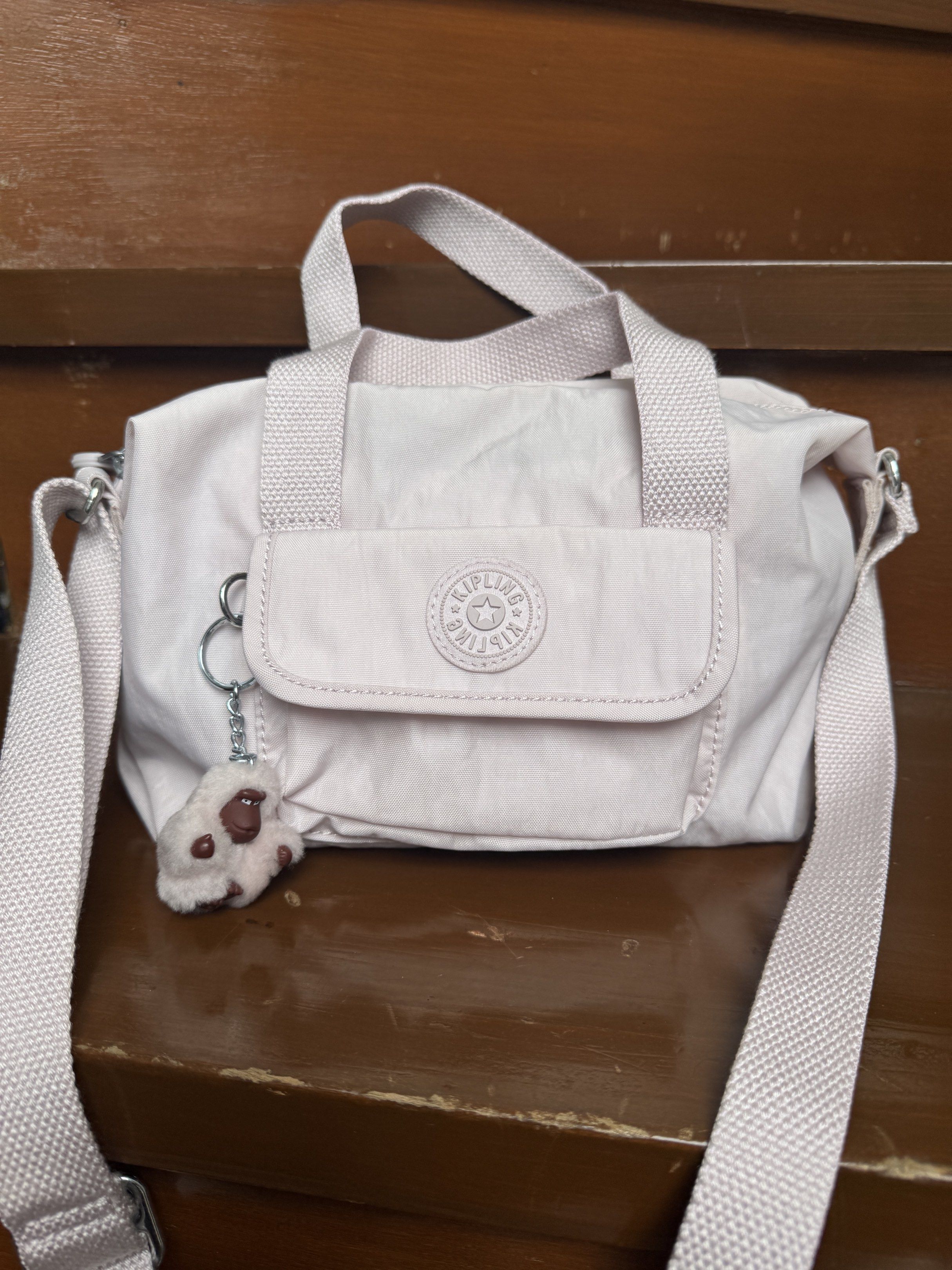 Kipling Brynn Serene Lavender Crossbody Bag (Category: Cross-body Bags) located in Paranaque City. Submitted by kenoh07 (Listing ID 1418185205, Image 1). Description: Kipling Brynn Serene Lavender crossbody bag. Features a light purple color with a detachable strap and top handles. Includes a small front pocket and a Kipling monkey keychain. The interior has a white lining with a small pocket. The bag has a zipper closure and Kipling branding on 