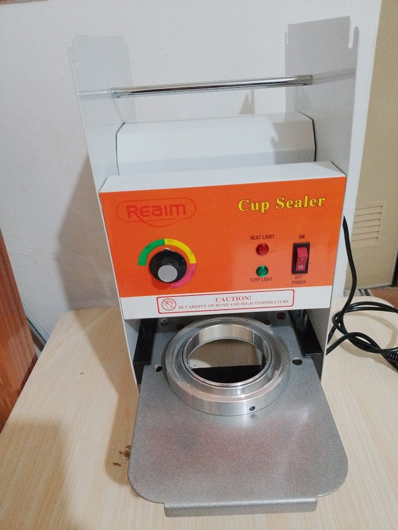 Realim Cup Sealer (Category: Other Kitchen Appliances) located in Manila. Submitted by mystictiger-1ff2 (Listing ID 1418104376, Image 1). Description: Realim cup sealer is perfect for sealing cups. It features an orange control panel with a heat limit indicator and power switch. The machine includes a sealing mechanism and comes with additional orange felt pieces and metal rods. It has a power cord and is designed for efficient cup sealing..
