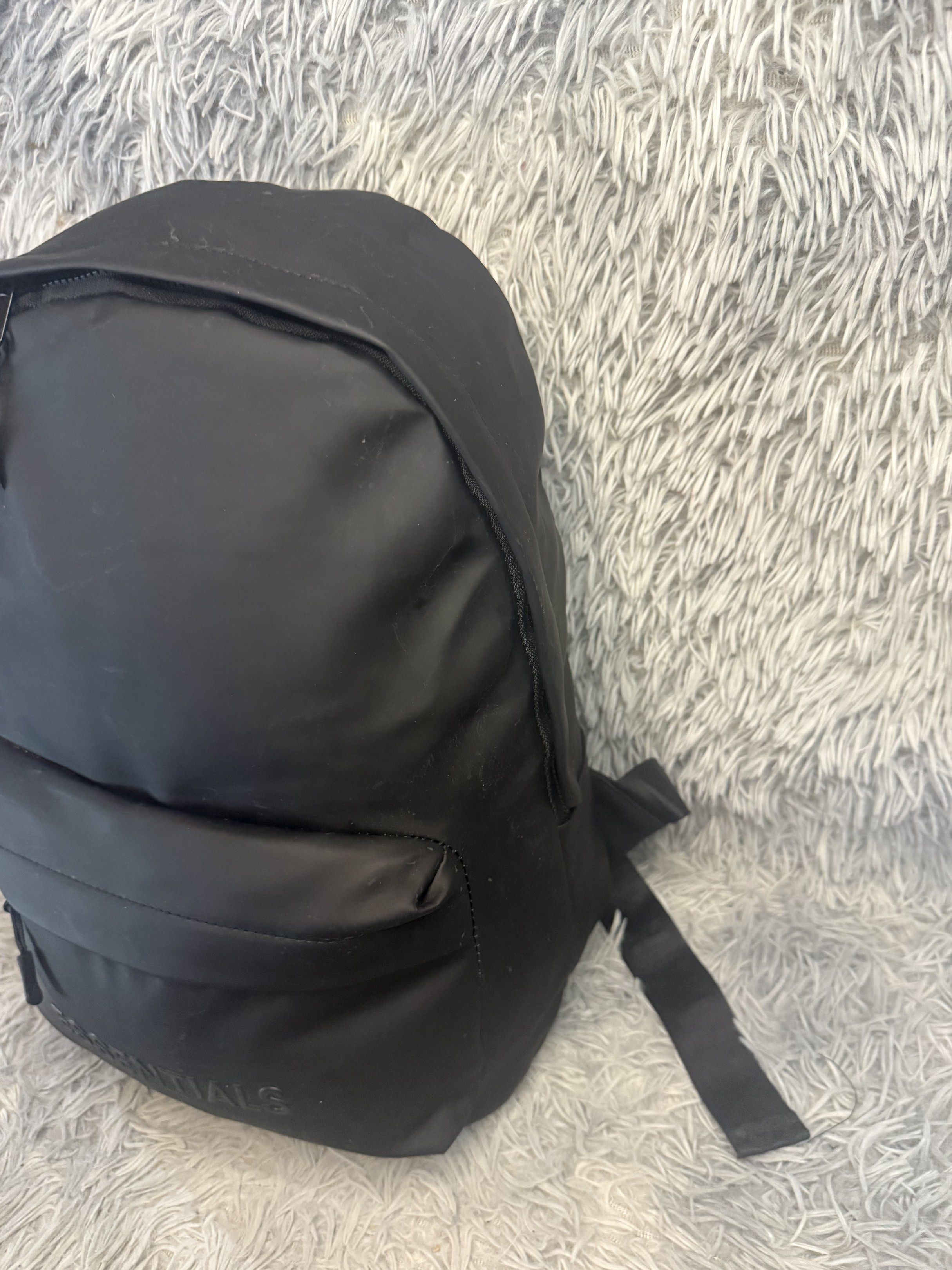 Fear of God Essentials Backpack Black (Category: Announcements) located in Davao. Submitted by Noel27 (Listing ID 1418300510, Image 3). Description: Black backpack with the Essentials logo embossed on the front. Features two zippered compartments and adjustable shoulder straps with Essentials branding. The interior has a small pocket and a tag with the Fear of God Essentials logo. The backpack is made of a smooth, possibly water-resistant materia