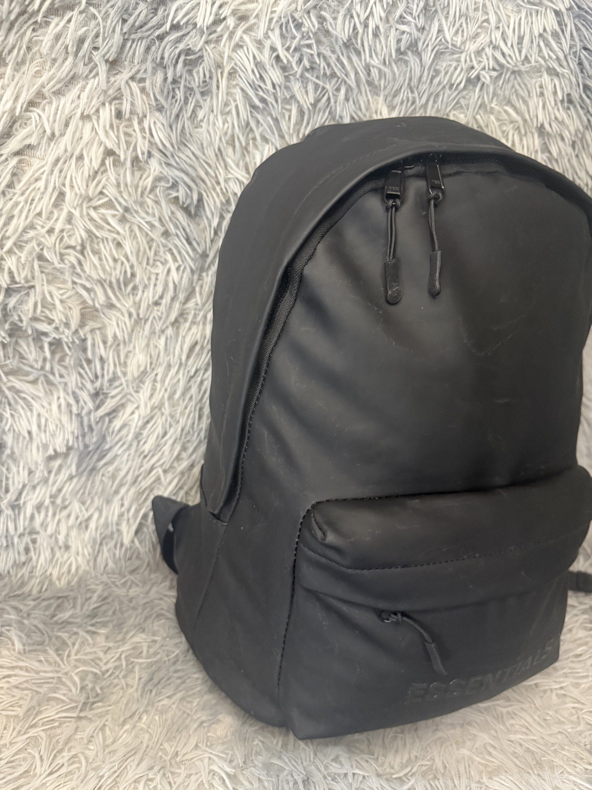 Fear of God Essentials Backpack Black (Category: Announcements) located in Davao. Submitted by Noel27 (Listing ID 1418300510, Image 2). Description: Black backpack with the Essentials logo embossed on the front. Features two zippered compartments and adjustable shoulder straps with Essentials branding. The interior has a small pocket and a tag with the Fear of God Essentials logo. The backpack is made of a smooth, possibly water-resistant materia