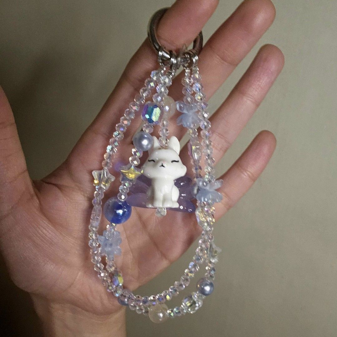 Cute Nine-tailed Fox phone/bag charm (Category: Charms) located in Las Pinas. Submitted by klya00 (Listing ID 1418451038, Image 1). Description: Adorable keychain features a white and blue Nine-tailed Fox figurine surrounded by iridescent beads, blue beads, and star-shaped charms. It also includes a phone tab to use as a phone charm. Perfect for adding a touch of charm to your phone or bag..