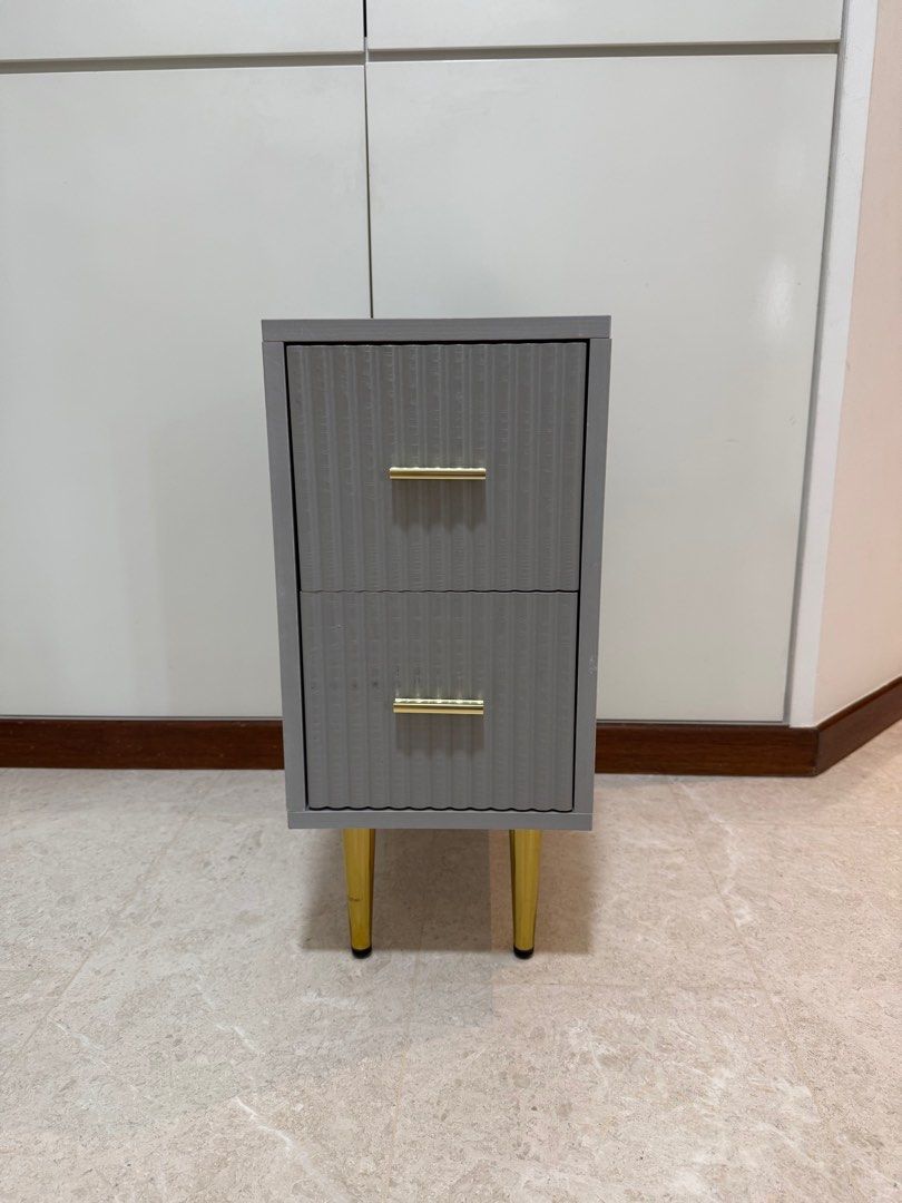 Bedside Table (Category: Tables & Sets) located in Singapore. Submitted by infinity80 (Listing ID 1413241405, Image 1). Description: - Bedside Table with two drawers - Dimensions: 25cm x 40 cm x 55cm (L x W x H) - Textured front design - Sophisticated grey finish - Gold-colored handles and legs - Perfect for bedrooms or living areas.