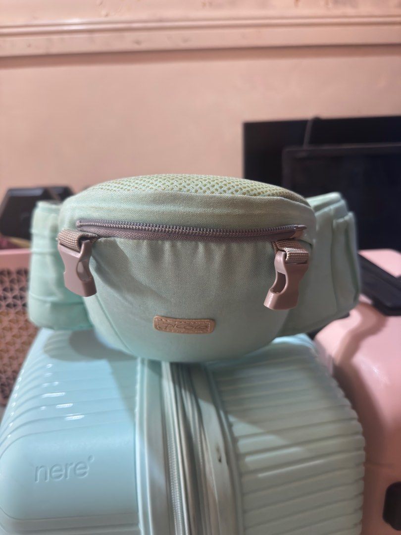 Picolo Baby Carrier (Category: Carriers & Slings) located in Makati City. Submitted by ms_gc01052003 (Listing ID 1413479603, Image 1). Description: - Mint green waist bag - Mesh top - Zippered main compartment - Side pockets - Brown logo patch - Brown buckles - Sits on a light blue suitcase.