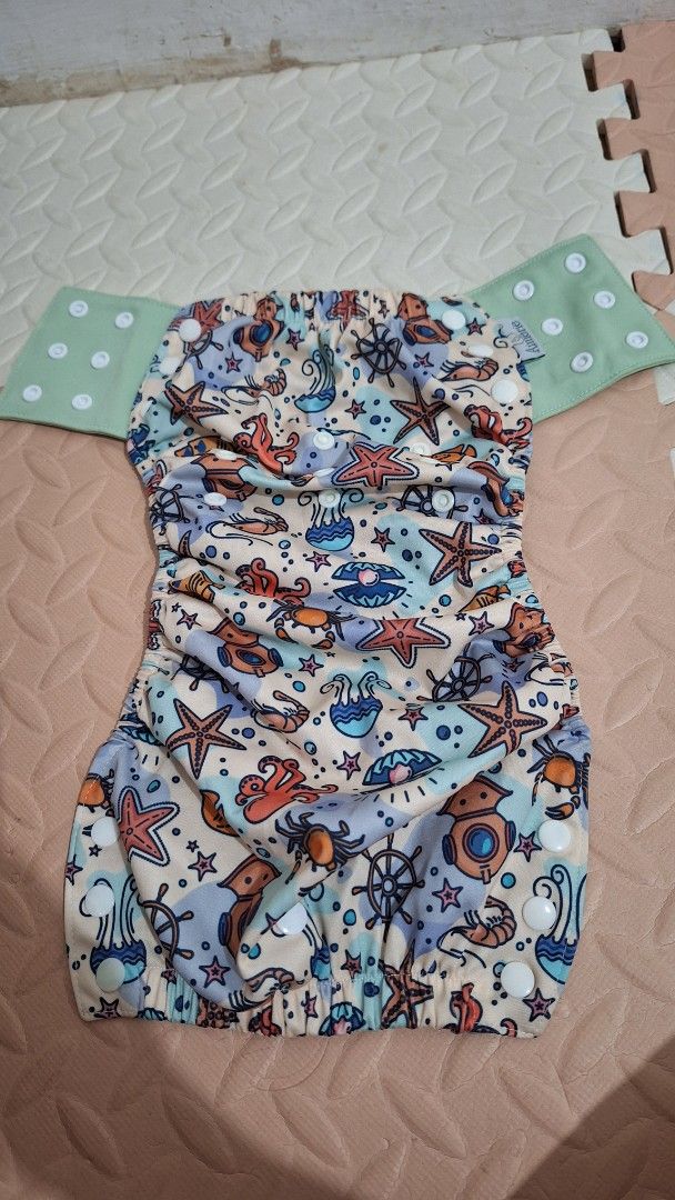 Aimerie Cloth Diaper (Category: Diapers & Baby Wipes) located in Cavite. Submitted by Tanya_Closet (Listing ID 1414106466, Image 2). Description: - Aimerie cloth diaper with a colorful sea creature design. - Features snap closures for easy adjustments. - Includes a separate green snap-on piece. - Designed with a white inner lining. - Has a stylish green trim. - Reusable and washable, making it an eco-friendly choice..