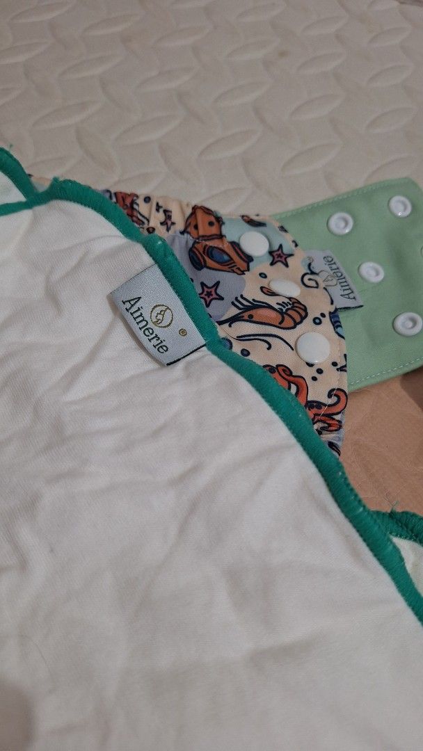 Aimerie Cloth Diaper (Category: Diapers & Baby Wipes) located in Cavite. Submitted by Tanya_Closet (Listing ID 1414106466, Image 1). Description: - Aimerie cloth diaper with a colorful sea creature design. - Features snap closures for easy adjustments. - Includes a separate green snap-on piece. - Designed with a white inner lining. - Has a stylish green trim. - Reusable and washable, making it an eco-friendly choice..