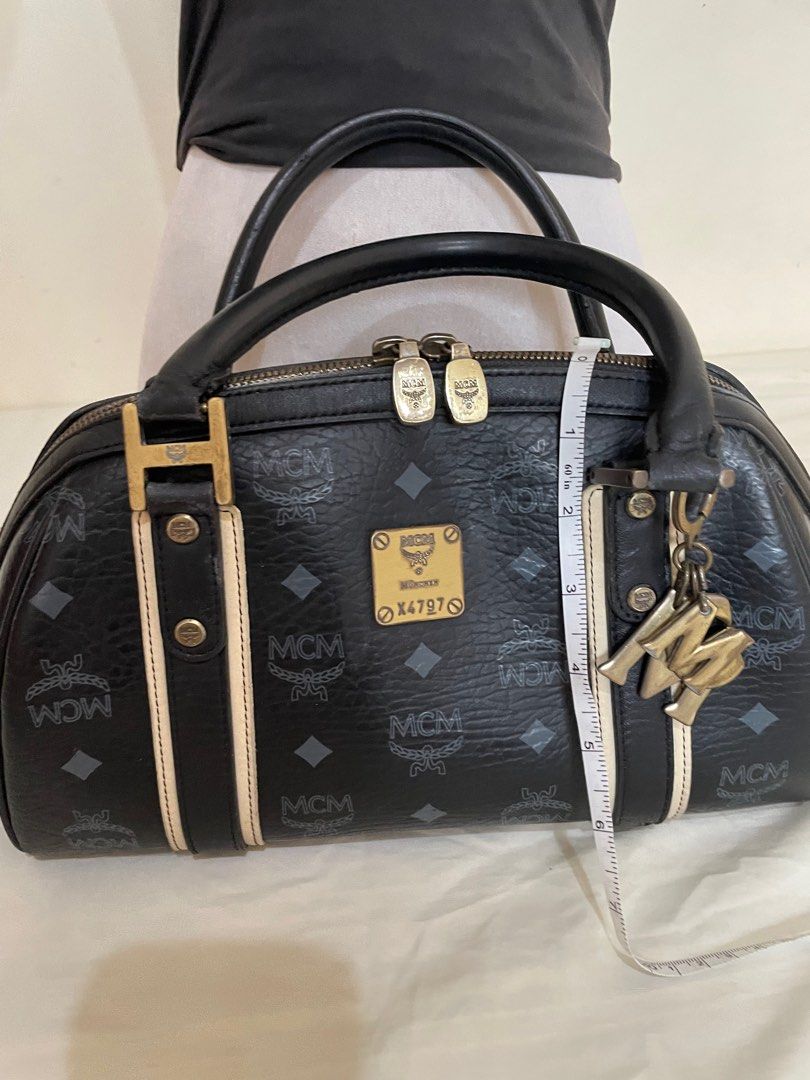 MCM Boston Bag (Category: Luxury Bags & Wallets) located in Tanauan. Submitted by airese (Listing ID 1414623347, Image 1). Description: Black MCM Boston bag with the brand's logo pattern. Features dual top handles, a zip closure, and a metal logo plate. Includes a detachable charm. The bag has a black interior with multiple compartments. The hardware shows some signs of wear. The bag has a beige stripe detail on the front and back..