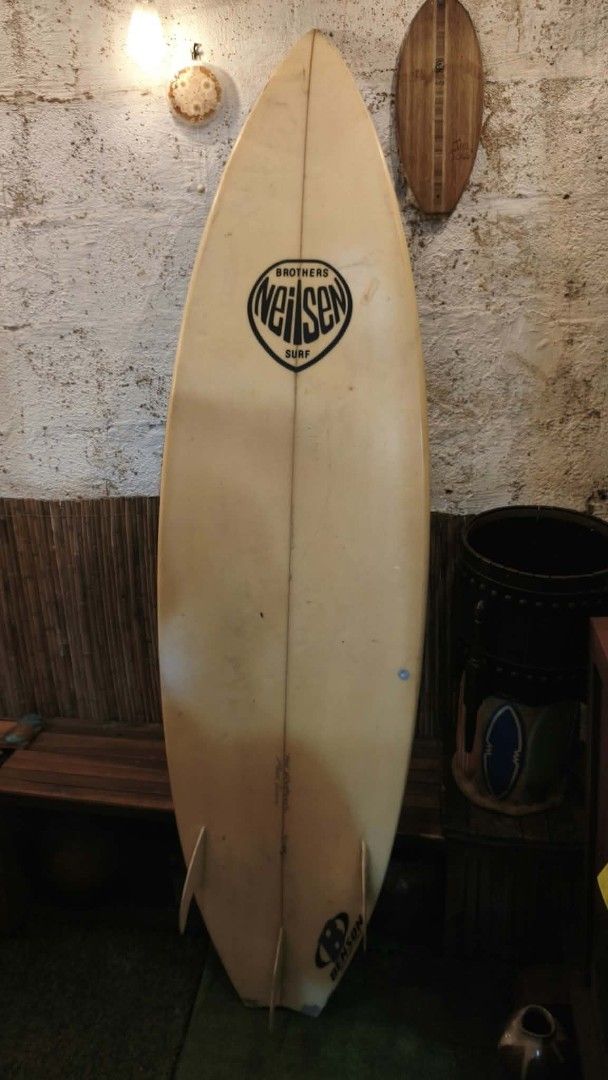 Brothers Neilsen Surf Surfboard (Category: Swim Trunks & Board Shorts) located in Manila. Submitted by glydelmalu86312 (Listing ID 1465320330, Image 2). Description: - Brand/Model: Brothers Neilsen surf board with Benson traction pad - Design: Classic white finish with central wooden stringer from nose to tail - Setup: Tri-fin configuration - Condition: Well-loved with wax residue, minor dings near the nose, a repaired patch near the logo, and a 